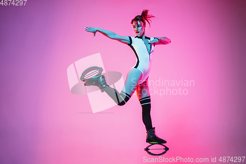 Image of Beautiful redhead woman in a white sportswear jumping in a kangoo jumps shoes isolated on gradient studio background in neon light