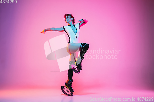 Image of Beautiful redhead woman in a white sportswear jumping in a kangoo jumps shoes isolated on gradient studio background in neon light