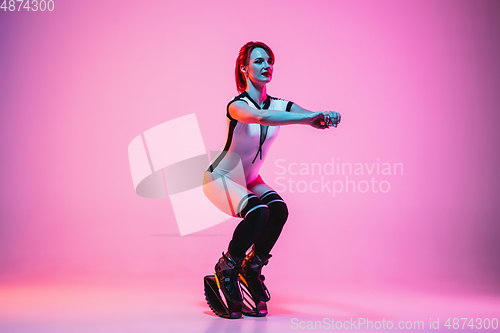 Image of Beautiful redhead woman in a white sportswear jumping in a kangoo jumps shoes isolated on gradient studio background in neon light