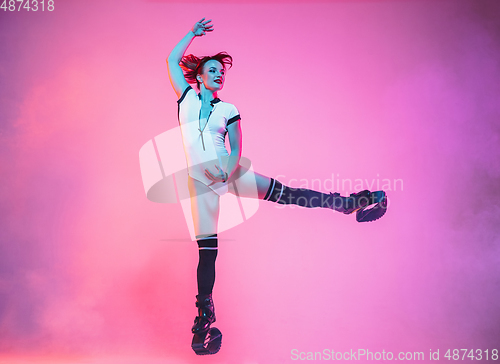 Image of Beautiful redhead woman in a white sportswear jumping in a kangoo jumps shoes isolated on gradient studio background in neon light