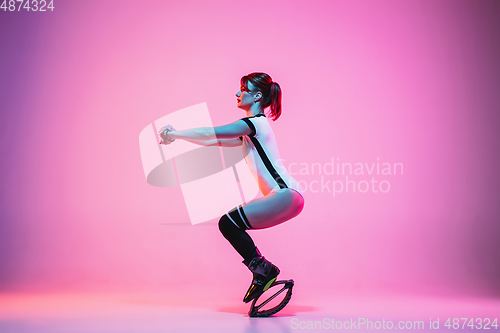 Image of Beautiful redhead woman in a white sportswear jumping in a kangoo jumps shoes isolated on gradient studio background in neon light