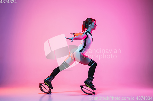 Image of Beautiful redhead woman in a white sportswear jumping in a kangoo jumps shoes isolated on gradient studio background in neon light