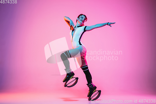 Image of Beautiful redhead woman in a white sportswear jumping in a kangoo jumps shoes isolated on gradient studio background in neon light