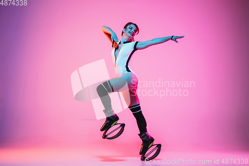 Image of Beautiful redhead woman in a white sportswear jumping in a kangoo jumps shoes isolated on gradient studio background in neon light