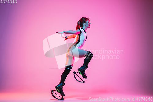 Image of Beautiful redhead woman in a white sportswear jumping in a kangoo jumps shoes isolated on gradient studio background in neon light