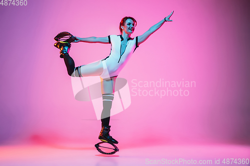 Image of Beautiful redhead woman in a white sportswear jumping in a kangoo jumps shoes isolated on gradient studio background in neon light