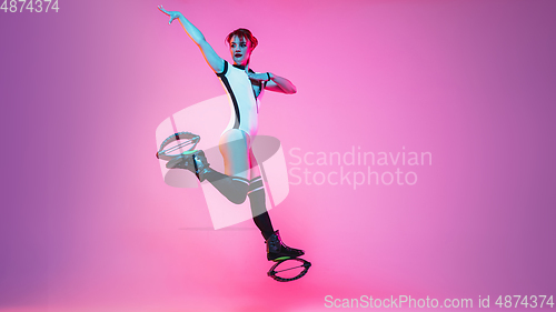 Image of Beautiful redhead woman in a white sportswear jumping in a kangoo jumps shoes isolated on gradient studio background in neon light
