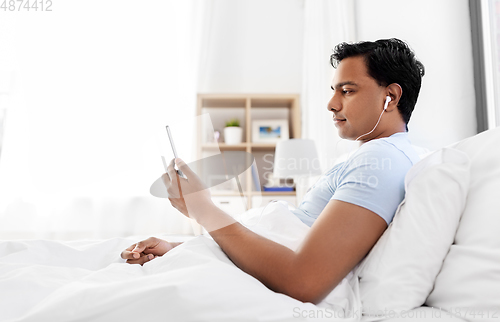 Image of indian man in earphones with phone in bed at home