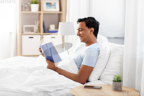 Image of happy indian man reading book in bed at home