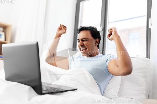 Image of happy indian man with laptop in bed at home