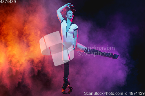 Image of Beautiful redhead woman in a white sportswear jumping in a kangoo jumps shoes isolated on gradient studio background in neon lighted smoke