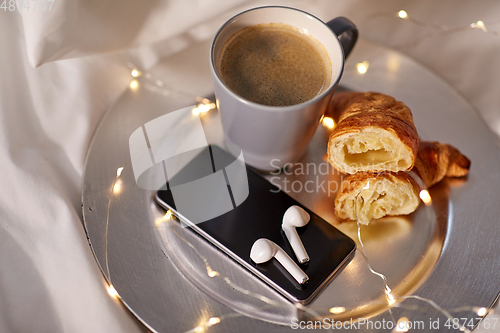 Image of smartphone, earphones, coffee and croissant in bed