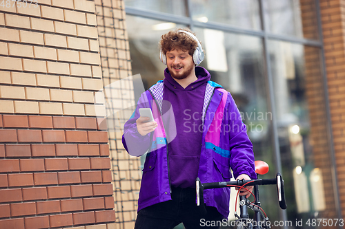 Image of Handsome young man using mobile phone and headphones while standing near his bicycle beside him