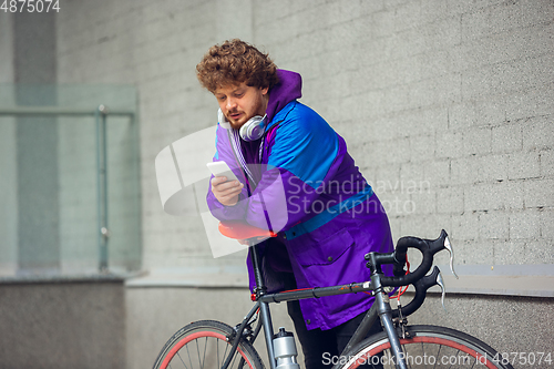 Image of Handsome young man using mobile phone and headphones while standing near his bicycle beside him