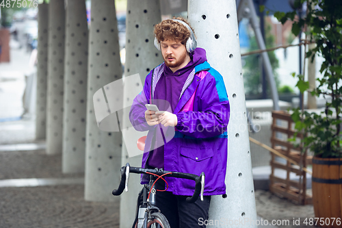 Image of Handsome young man using mobile phone and headphones while standing near his bicycle beside him