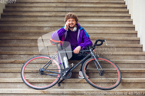 Image of Handsome young man using mobile phone and headphones while sitting near his bicycle beside him