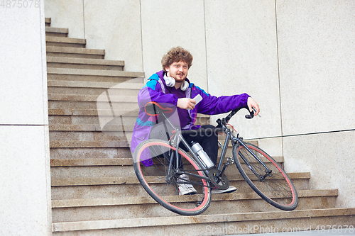 Image of Handsome young man using mobile phone and headphones while sitting near his bicycle beside him