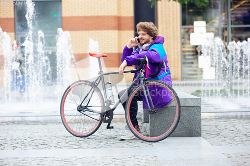 Image of Handsome young man using mobile phone and headphones while sitting near his bicycle beside him