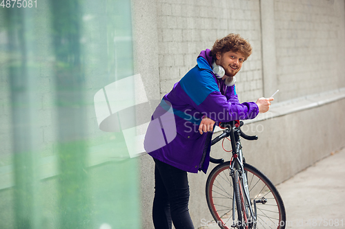 Image of Handsome young man using mobile phone and headphones while standing near his bicycle beside him