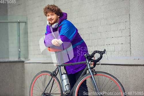 Image of Handsome young man using mobile phone and headphones while standing near his bicycle beside him