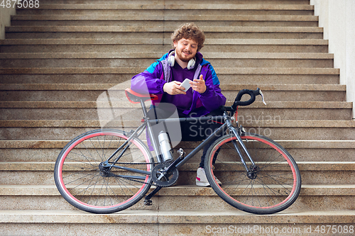 Image of Handsome young man using mobile phone and headphones while sitting near his bicycle beside him