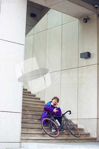 Image of Handsome young man using mobile phone and headphones while sitting near his bicycle beside him