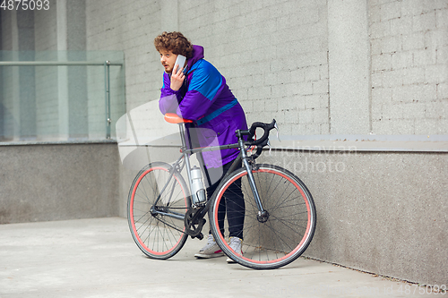 Image of Handsome young man using mobile phone and headphones while standing near his bicycle beside him