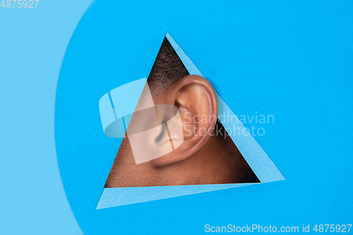 Image of Ear of african-american man peeking throught triangle in blue background