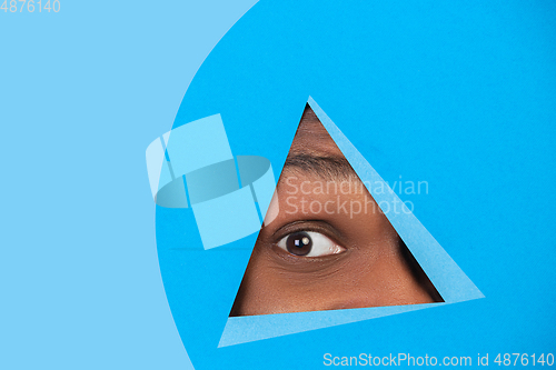 Image of Eye of african-american man peeking throught triangle in blue background