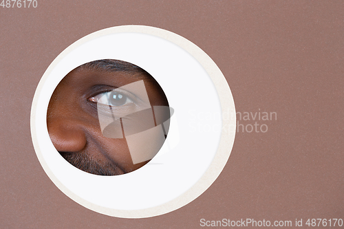 Image of Eye of smiling african-american man peeking throught circle in brown background