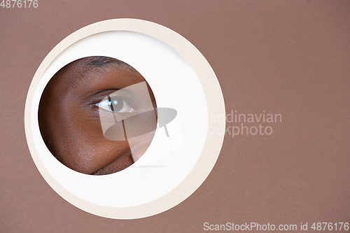 Image of Eye of smiling african-american man peeking throught circle in brown background