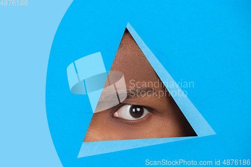 Image of Eye of african-american man peeking throught triangle in blue background