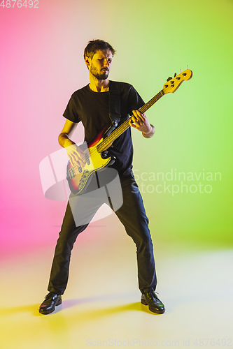 Image of Young caucasian musician playing bass guitar in neon light on pink-green background