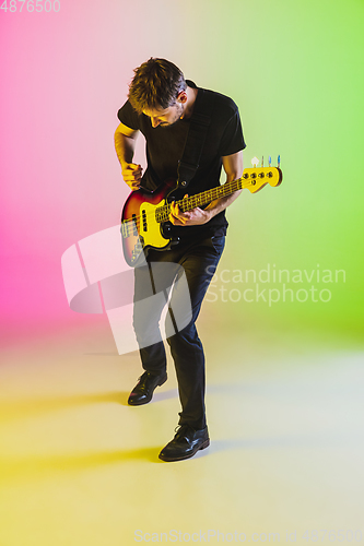 Image of Young caucasian musician playing bass guitar in neon light on pink-green background