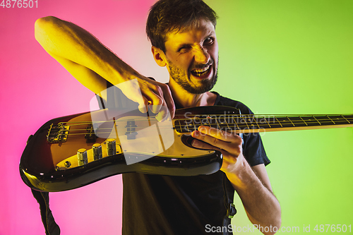 Image of Young caucasian musician playing bass guitar in neon light on pink-green background