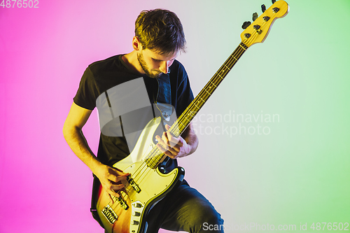 Image of Young caucasian musician playing bass guitar in neon light on pink-green background