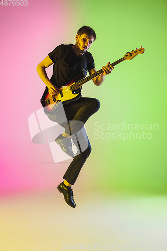 Image of Young caucasian musician playing bass guitar in neon light on pink-green background