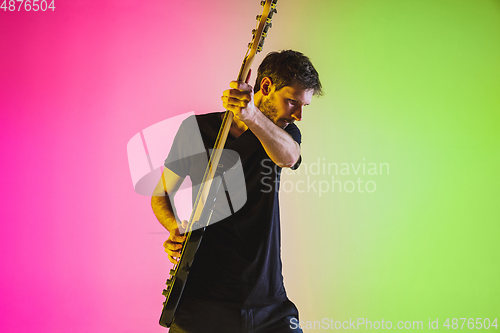 Image of Young caucasian musician playing bass guitar in neon light on pink-green background