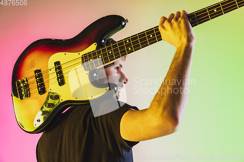 Image of Young caucasian musician playing bass guitar in neon light on pink-green background