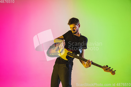 Image of Young caucasian musician playing bass guitar in neon light on pink-green background