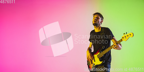 Image of Young caucasian musician playing bass guitar in neon light on pink-green background