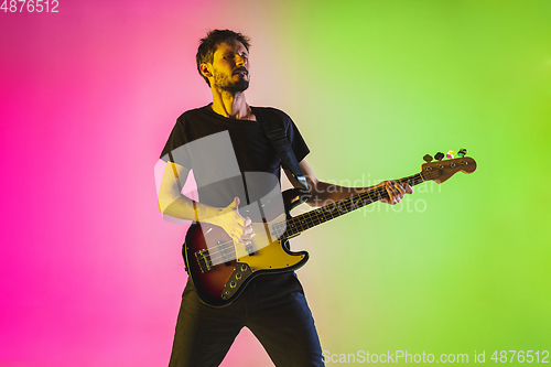 Image of Young caucasian musician playing bass guitar in neon light on pink-green background