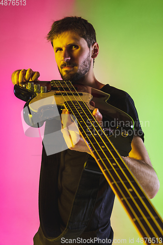 Image of Young caucasian musician playing bass guitar in neon light on pink-green background