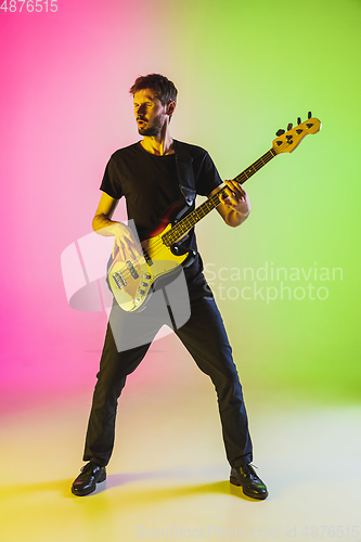 Image of Young caucasian musician playing bass guitar in neon light on pink-green background