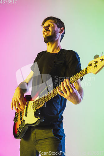 Image of Young caucasian musician playing bass guitar in neon light on pink-green background