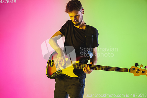 Image of Young caucasian musician playing bass guitar in neon light on pink-green background