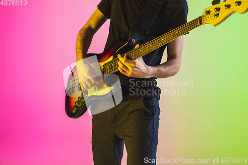 Image of Young caucasian musician playing bass guitar in neon light on pink-green background