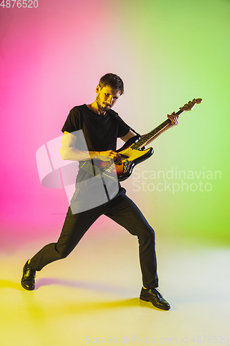 Image of Young caucasian musician playing bass guitar in neon light on pink-green background