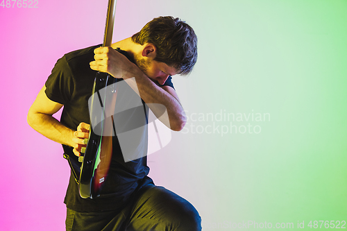 Image of Young caucasian musician playing bass guitar in neon light on pink-green background