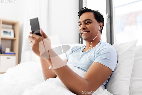 Image of indian man in earphones with phone in bed at home